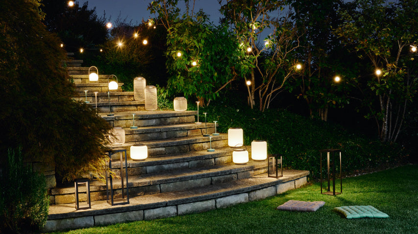 Beleuchtete Laternen und Lichterketten erhellen eine Gartensteintreppe am Abend, umgeben von Bäumen und Rasen.