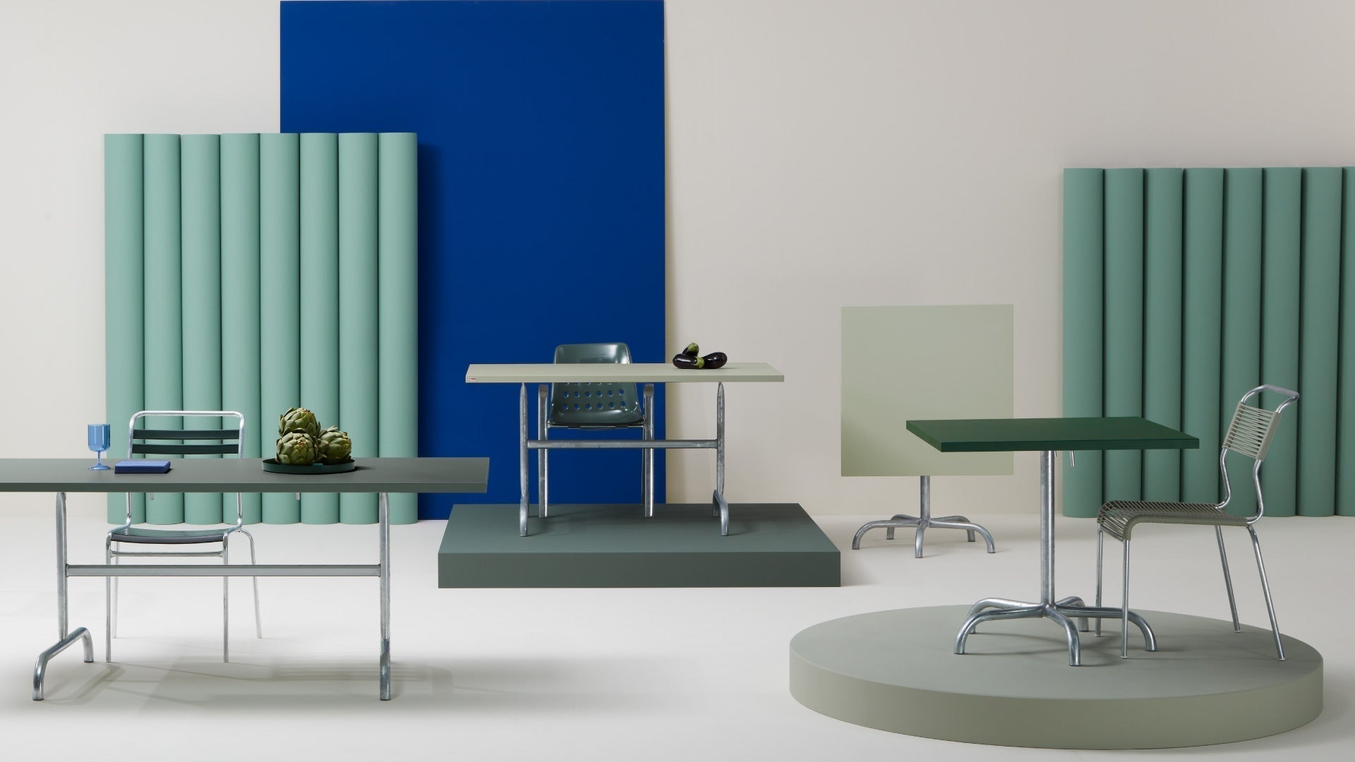 Verschiedene moderne Gartentische und -stühle in einer stilisierten Studio-Szene mit farbigen Paneelen im Hintergrund.