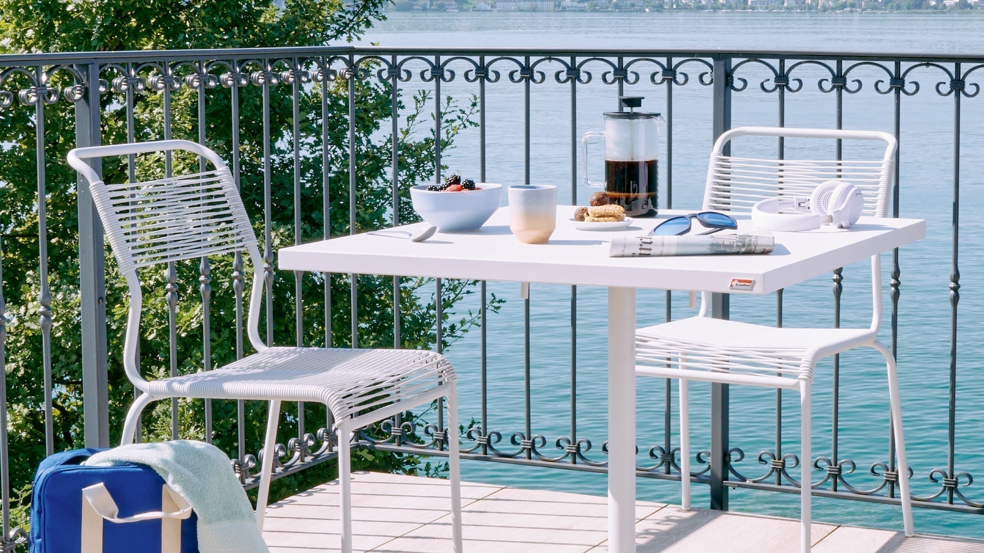 Gedeckter Balkontisch mit zwei weissen Gartenstühlen, Frühstücksutensilien und See im Hintergrund.