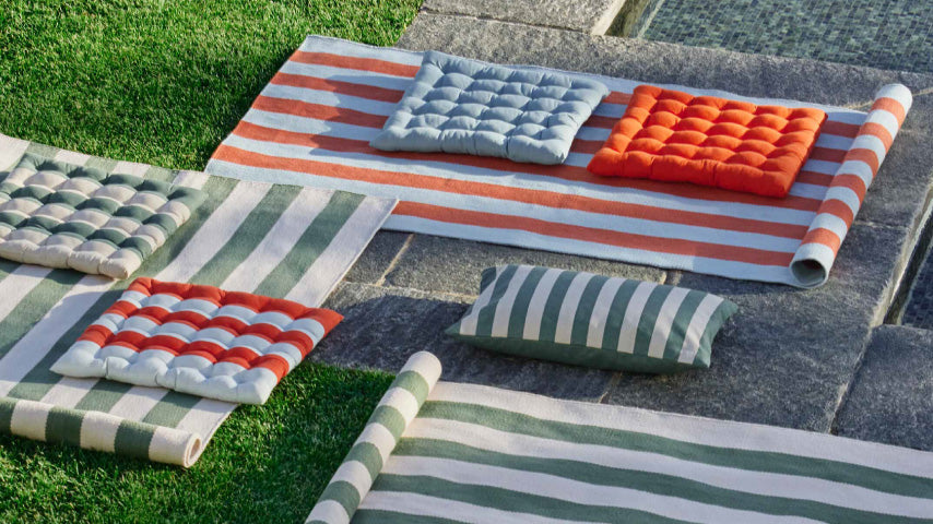 Gestreifte Outdoor-Teppiche und farbige Sitzkissen liegen auf Steinplatten und Rasen in der Sonne.