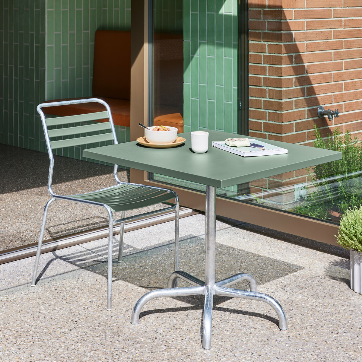 Grüner Tisch mit Metallstuhl auf einer Terrasse vor einer Glas- und Ziegelwand, darauf eine Schale, Tasse und ein Buch.