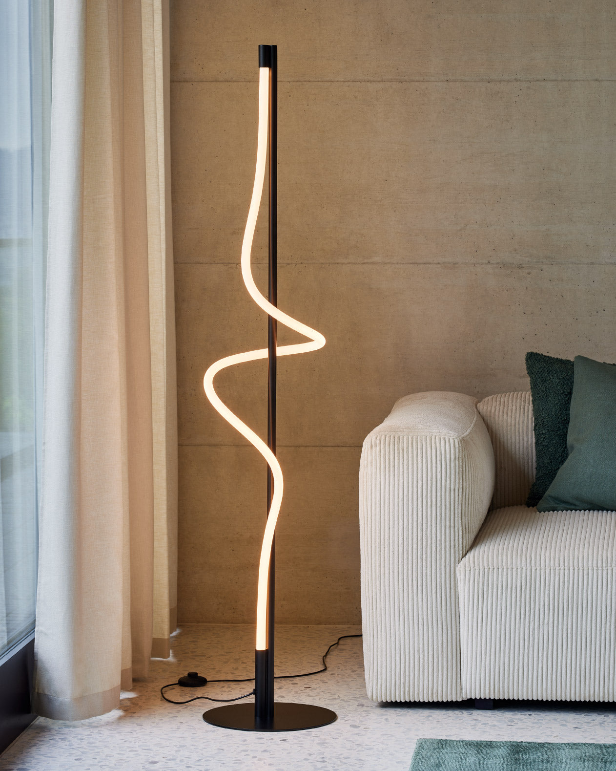 Schwarze Stehlampe mit geschwungenem LED-Lichtstreifen neben beigem Sofa und grüner Kissen.