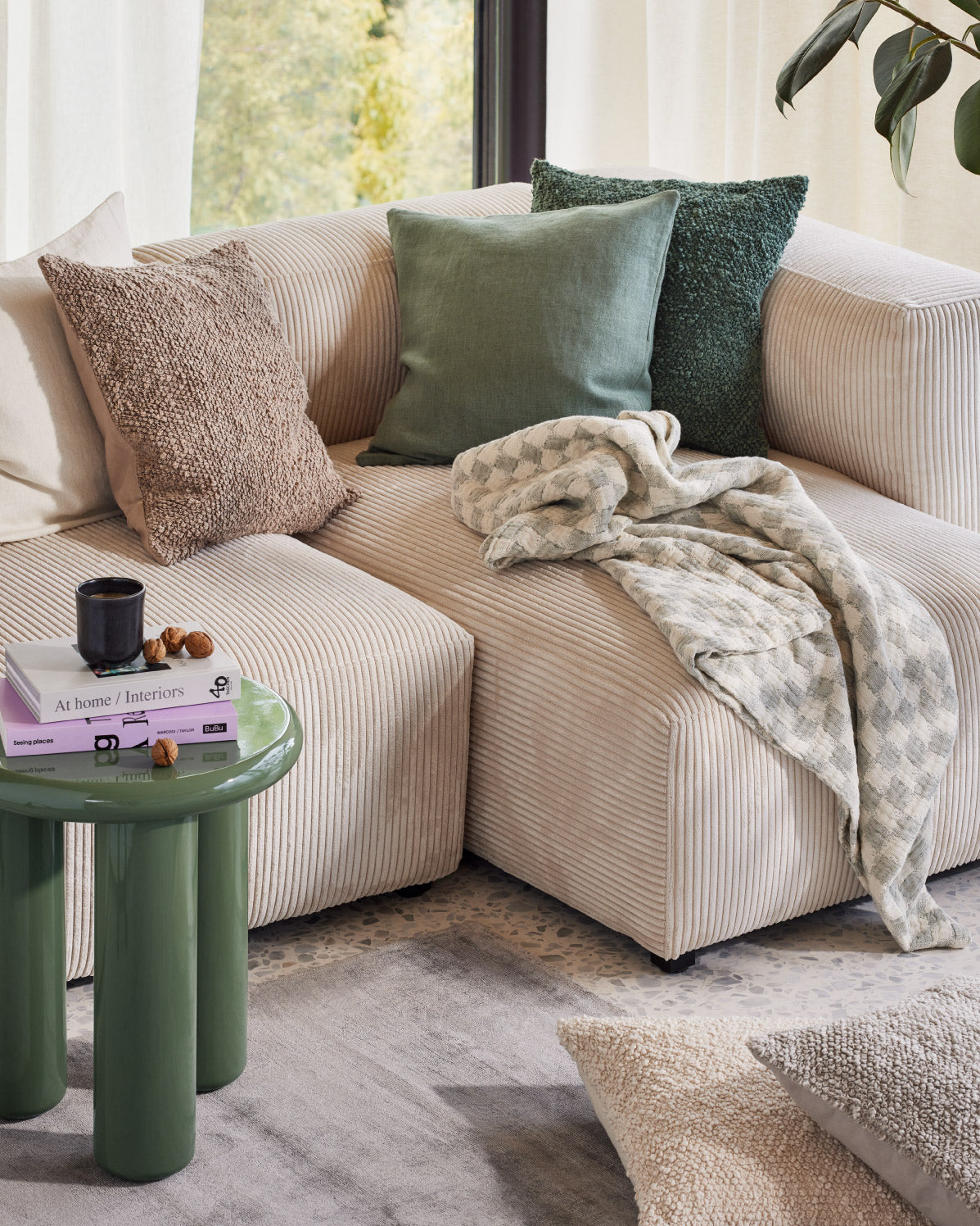 Beiges Sofa mit drei Kissen in Gruen- und Brauntoenen, darauf eine gemusterte Decke, daneben ein gruener Beistelltisch mit Buch und Tasse.