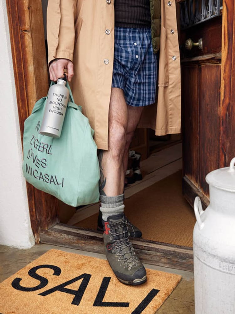 Personne en chaussures de randonnée et manteau avec un sac et une gourde sur un paillasson "Sali".