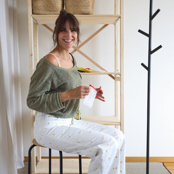 Person sitzt auf einem Hocker und hält Stoff und Schere, im Hintergrund ein Regal mit Körben.