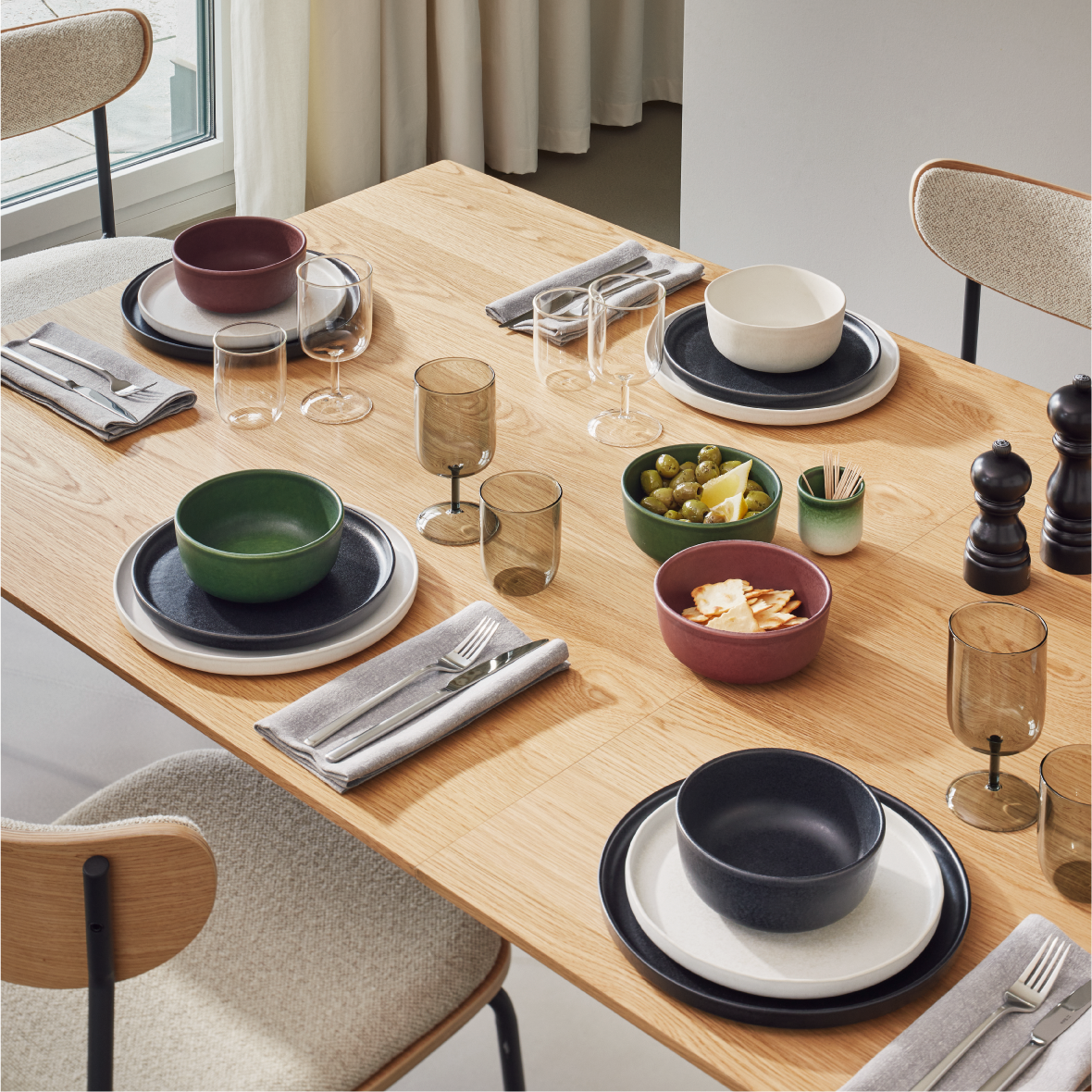 Holztisch mit modernem Geschirr, farbigen Schalen, Gläsern, Besteck und kleinen Snacks wie Oliven und Crackern.