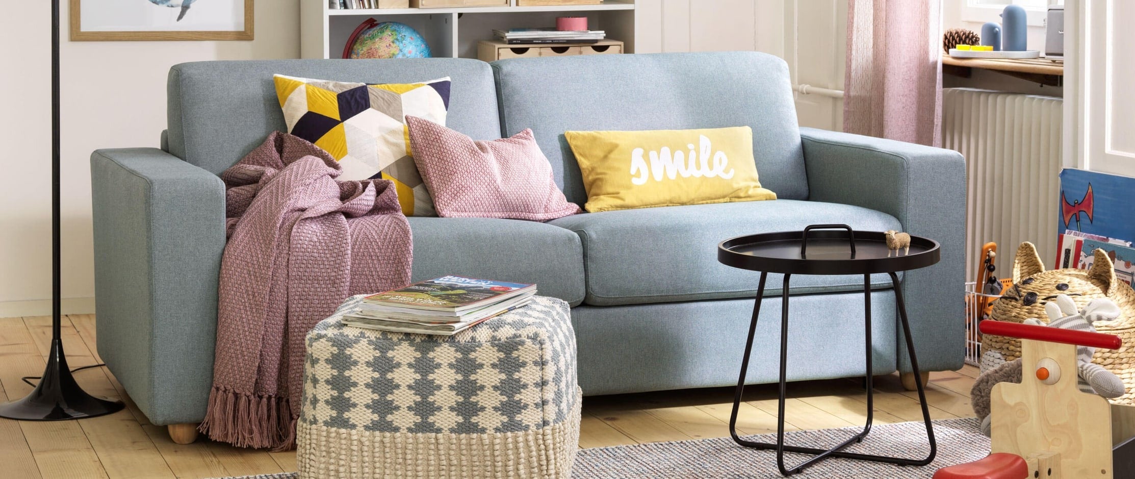 Ein Wohnzimmer, in welchem sich ein Sofa mit Kissen und einer Decke befindet und auf dem Boden Kinderspielzeug, wie Lego und ein Kindervelo liegen..jpg