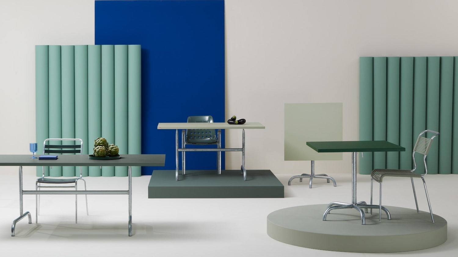 Diversi tavoli e sedie da giardino moderni in una scena stilizzata da studio con pannelli colorati sullo sfondo.