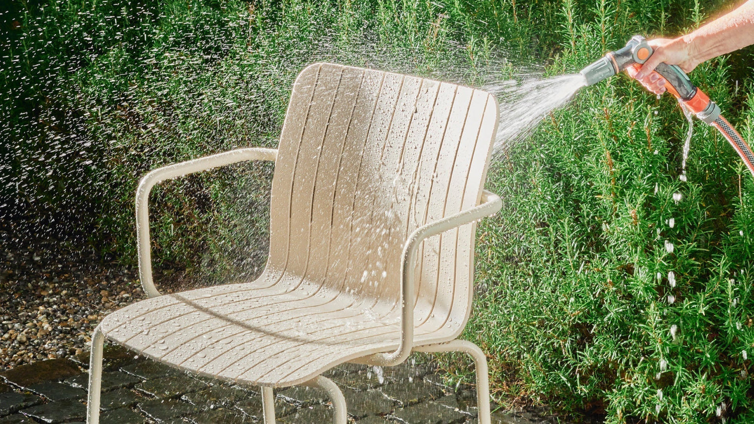 Ein Gartenstuhl wird im Freien mit einem Wasserschlauch abgespritzt, sodass Wasser über die Sitzfläche spritzt.