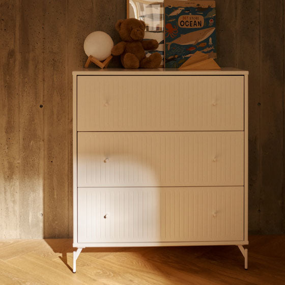 Weisse Kommode mit drei Schubladen, dekoriert mit Spielzeug und Büchern vor einer Betonwand.