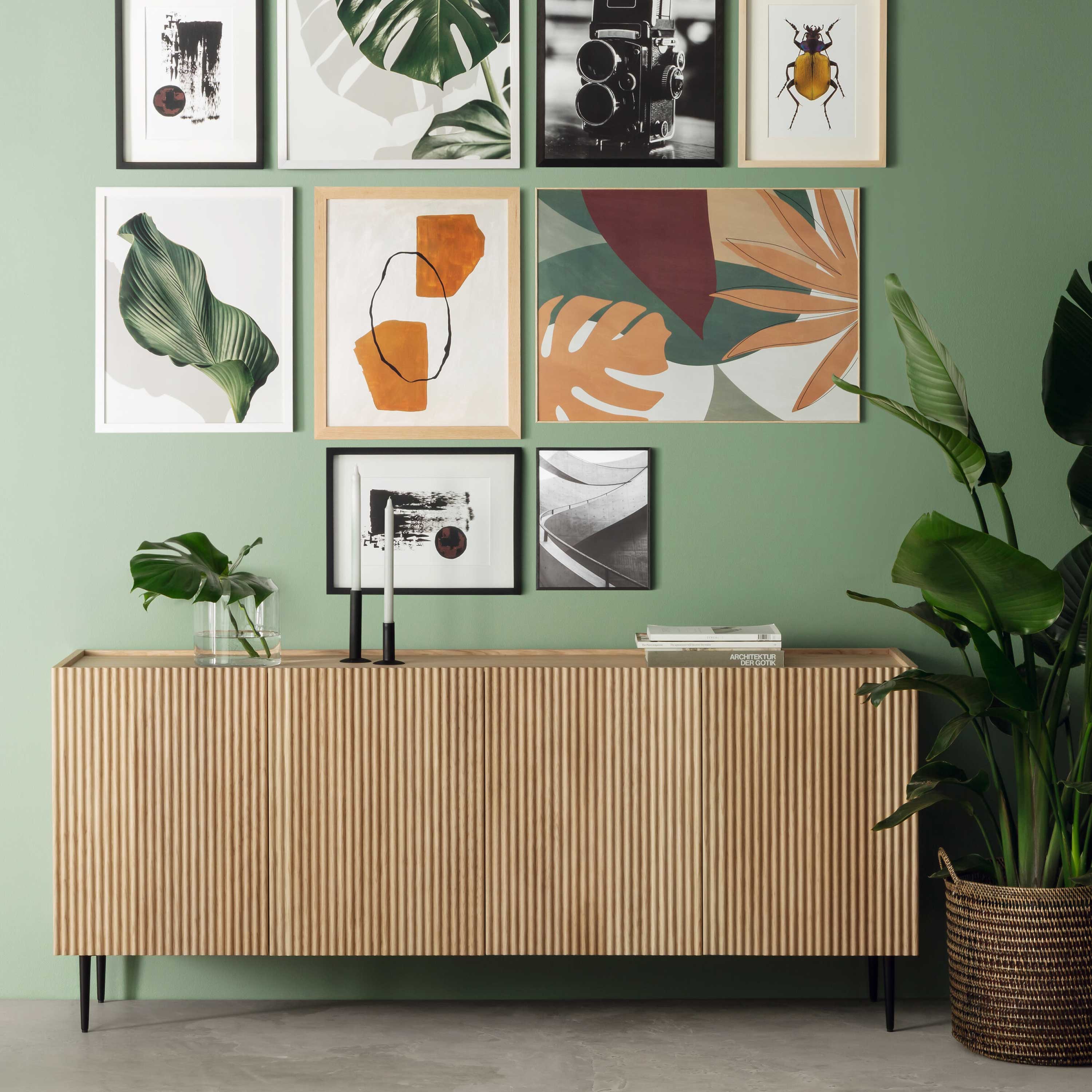 Holzsideboard vor grüner Wand mit dekorativer Bildergalerie und grossen Zimmerpflanzen.