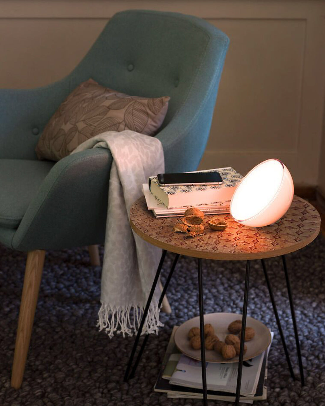 Blauer Sessel mit Kissen und heller Decke, daneben runder Holztisch mit Lampe, Büchern und Nüssen.