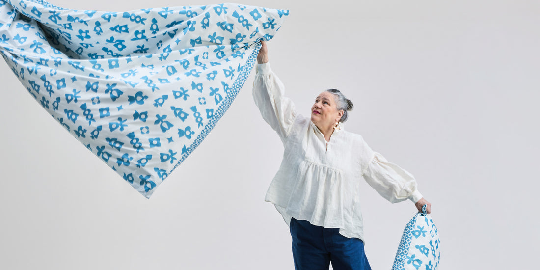 Sonnhild Kestler hält eine gemusterte Bettdecke in der Luft und trägt dazu passenden Kissenbezug in heller, minimalistischer Umgebung.