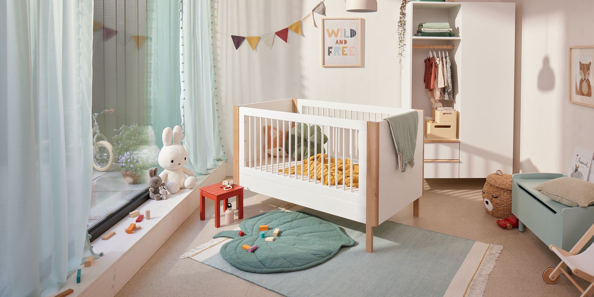 Helles Kinderzimmer mit weißem Gitterbett, bunten Wimpeln, offenem Kleiderschrank und Spielmatte in Blattform auf hellblauem Teppich.