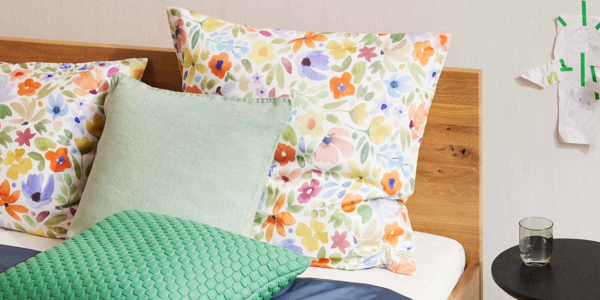 Bett mit floralen Kissen, mintgrünem Kissen und grüner Strukturdecke auf blauem Bettbezug.