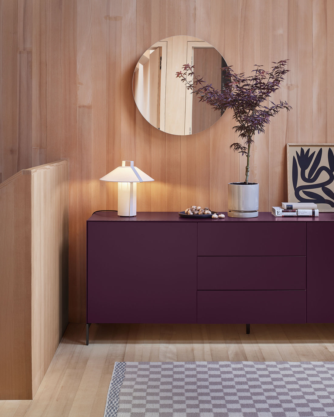 Modernes Wohnzimmer-Setup mit einem bordeauxfarbenen Sideboard, einer Tischlampe, einer Vase mit Zweigen und einem runden Wandspiegel vor einer Holzverkleidung.