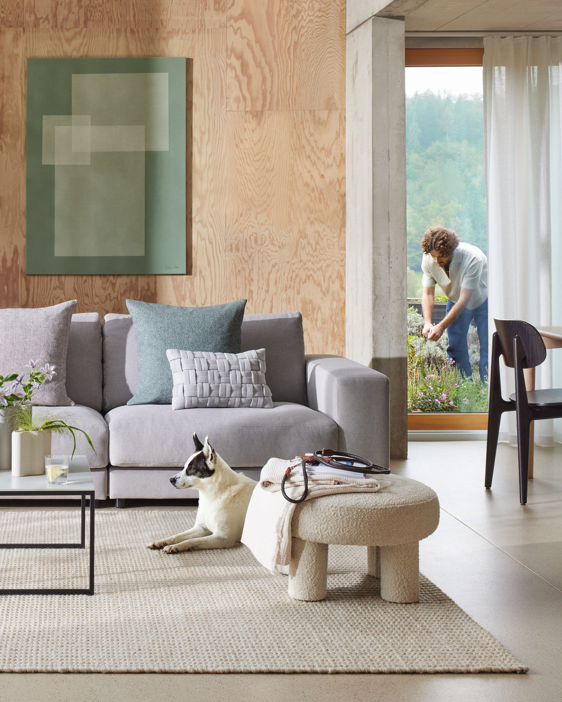 Modernes Wohnzimmer mit grauem Sofa, rundem Hocker und Hund im Vordergrund, während im Hintergrund eine Person durch ein offenes Fenster im Garten arbeitet.