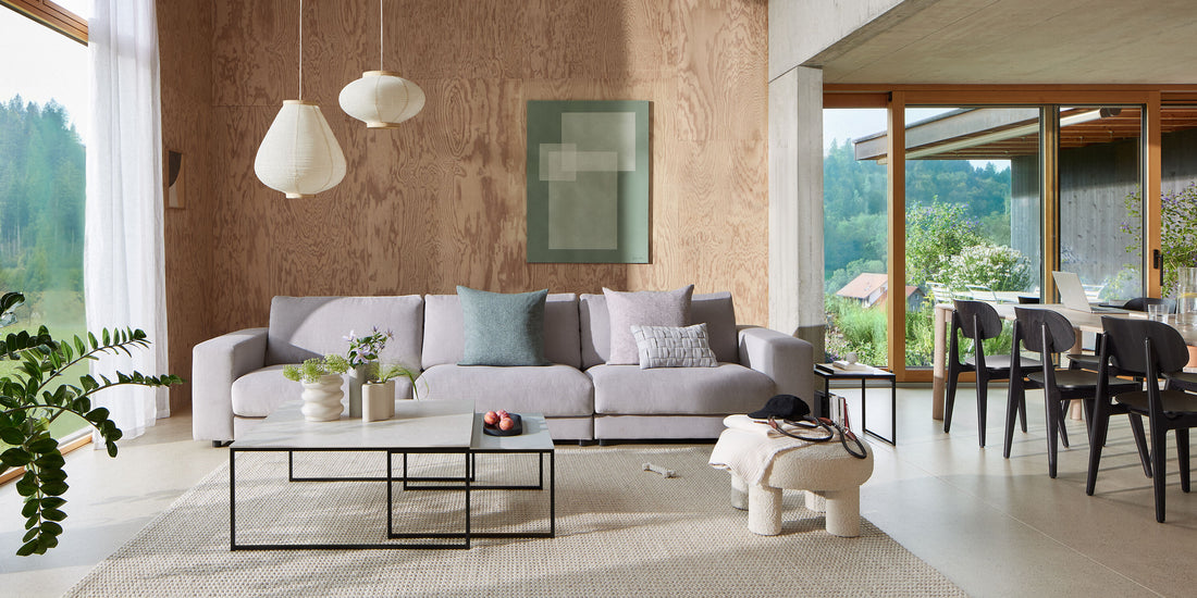Modernes Wohnzimmer mit hellgrauem Sofa, neutralen Holzwänden, grossen Fenstern mit Blick ins Grüne und minimalistischer Einrichtung.