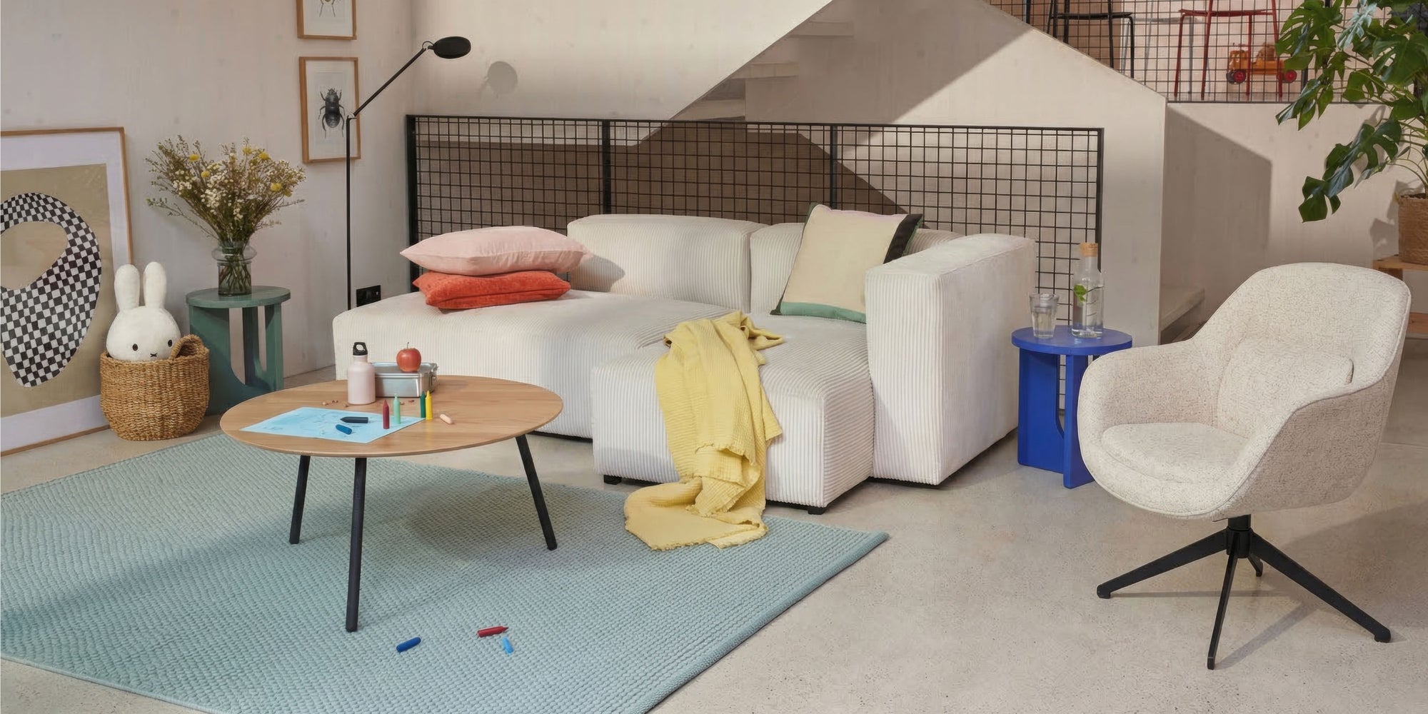 Modernes Wohnzimmer mit weissem Sofa, pastellfarbenen Kissen, rundem Holztisch und hellblauem Teppich unter einer Treppe.