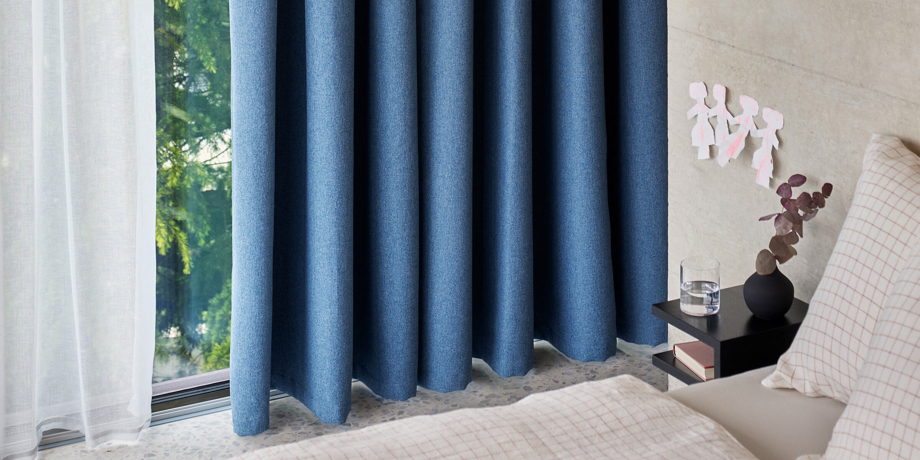 Bett neben einem Fenster mit weissen und blauen Vorhängen, kleine schwarze Ablage mit Glas und Deko.