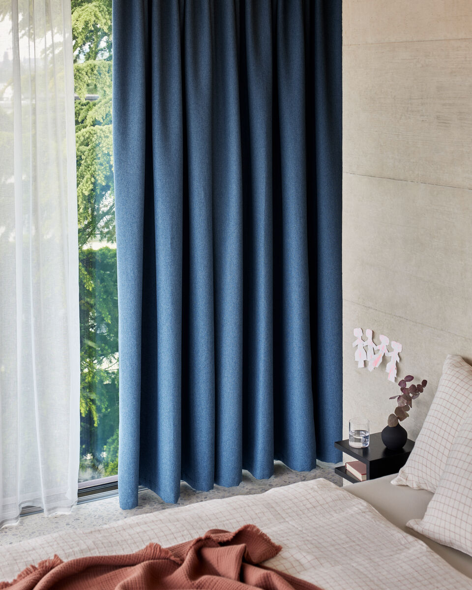 Schlafzimmer mit blauem Vorhang, weissem Bettwaesche-Set und brauner Decke auf dem Bett.