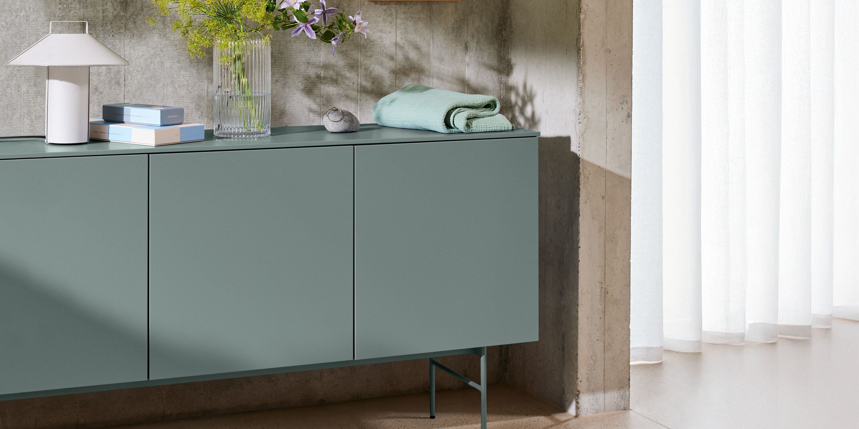 Modernes Sideboard in Salbeigrün mit Tischleuchte, Glasvase mit Blumen und dekorativen Wohnaccessoires.