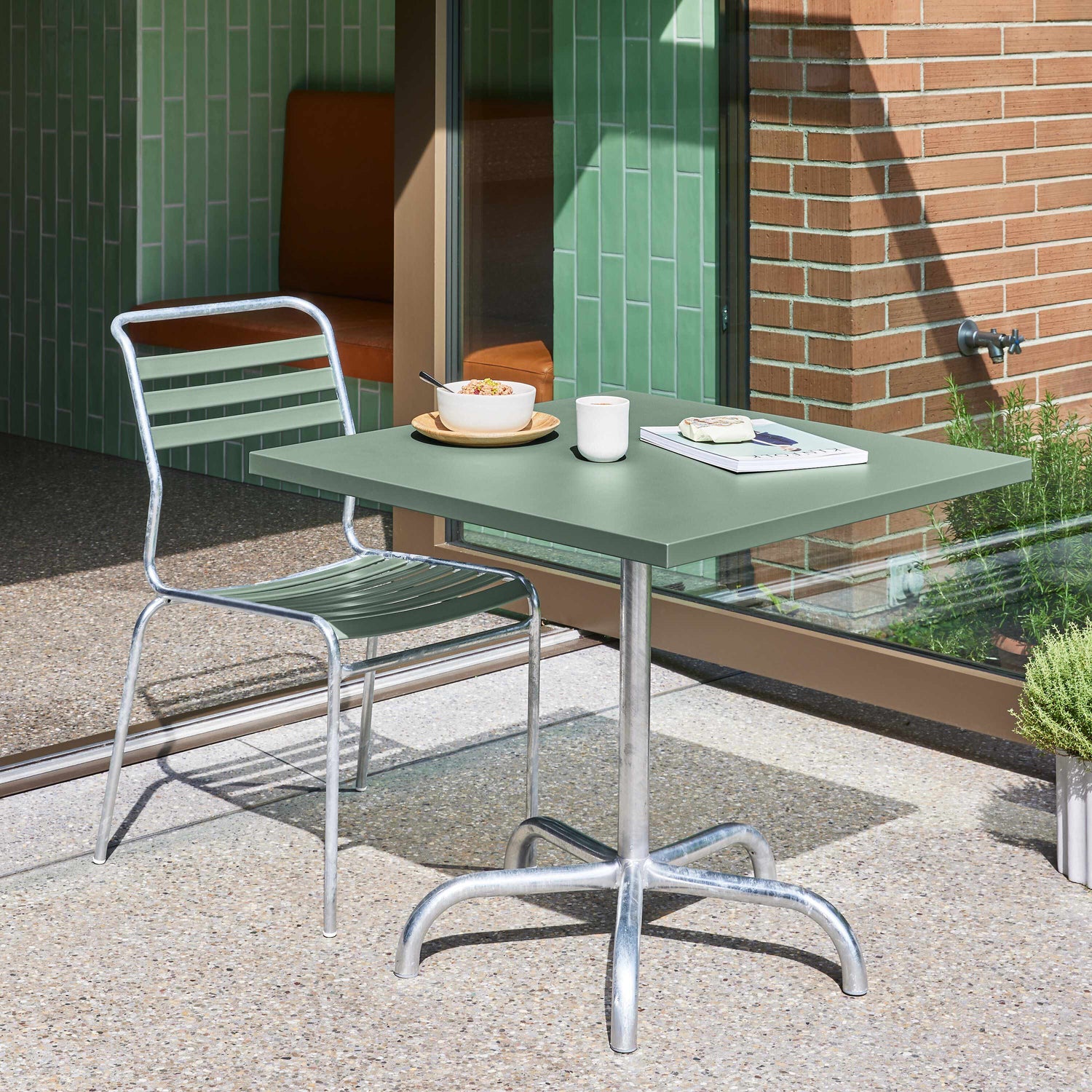 Kleiner Outdoor-Bereich mit grünem Metalltisch, passendem Stuhl und Frühstücksgeschirr vor einer Ziegel- und Glasfront.