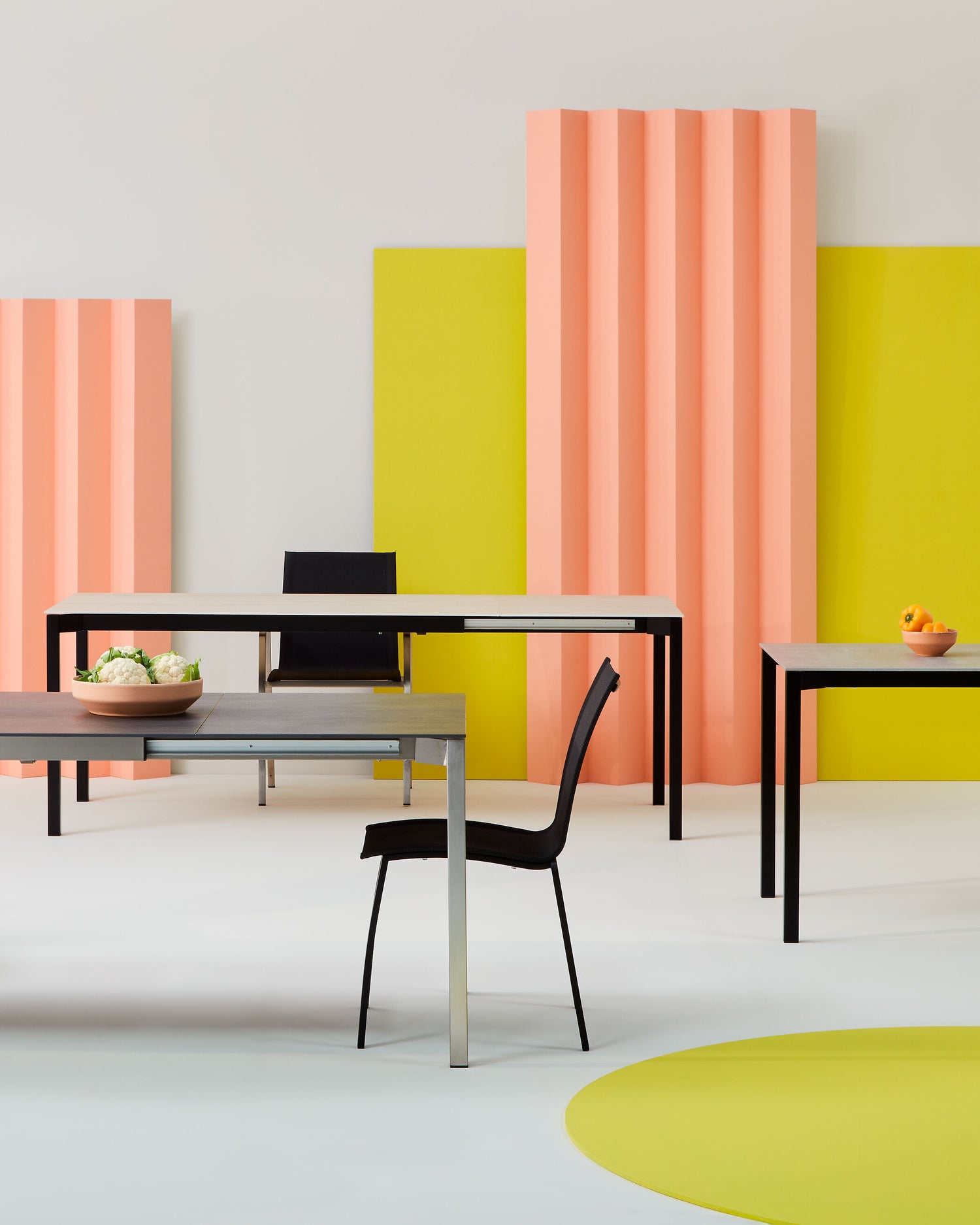 Modernes Interieur mit drei rechteckigen Tischen und schwarzen Stühlen vor einer Kulisse aus gelben und rosa Paneelen, dekoriert mit Schalen voller Obst und Blumen.