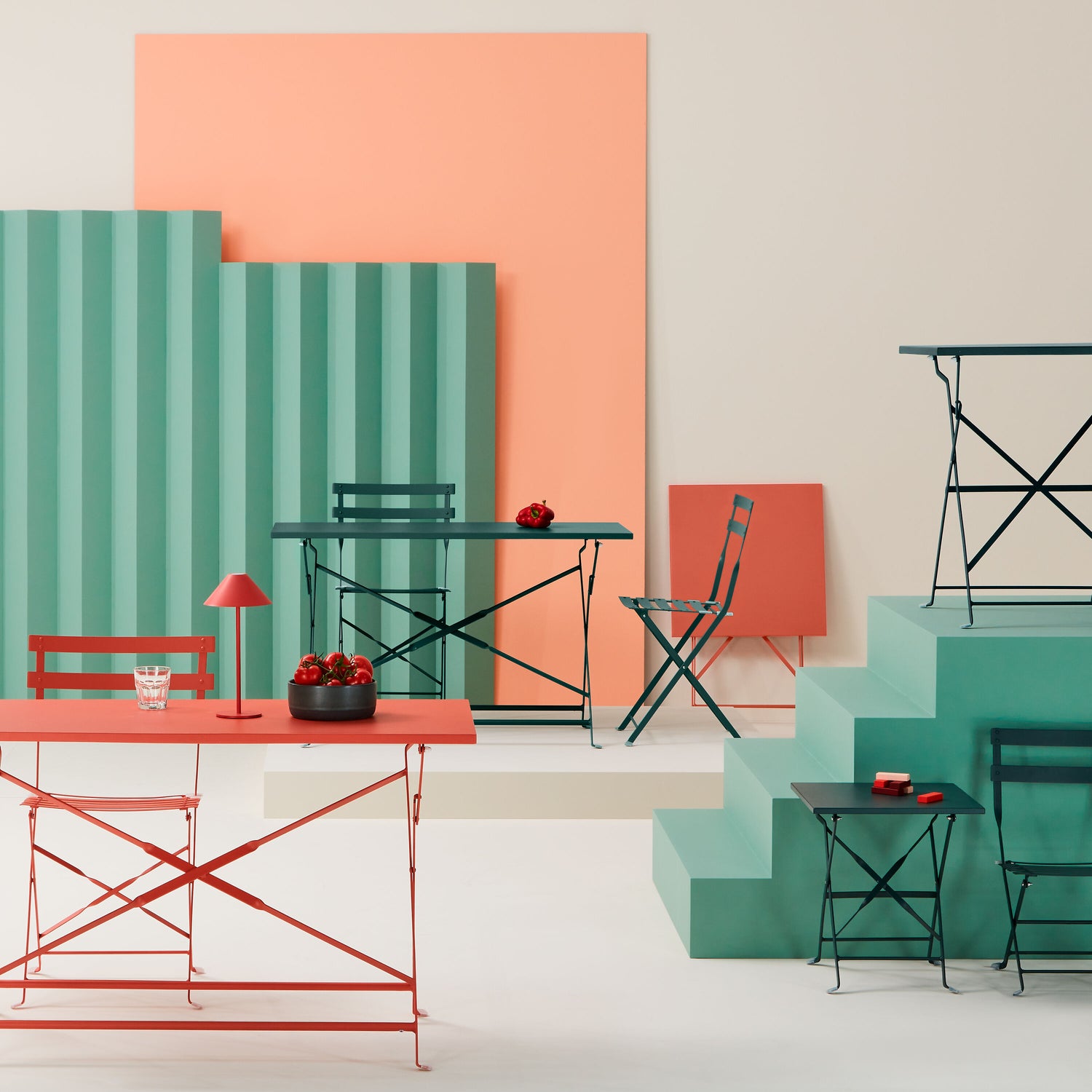 Farbige Outdoor‑Sitzgruppe mit klappbaren Metalltischen und -stühlen in Korall‑ und Grüntönen vor grafisch gestalteten Hintergrundelementen.