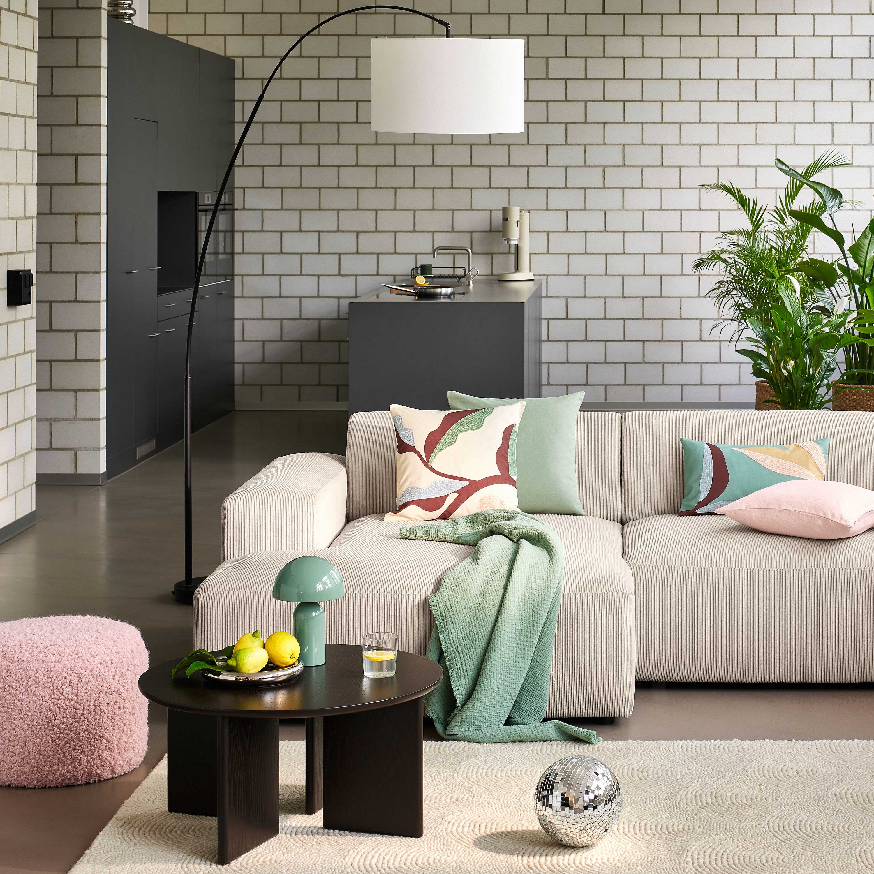 Modernes Wohnzimmer mit hellem Sofa, farbigen Kissen, Rundtisch mit Deko, grossem Bogenlampenlicht und offener Küche im Hintergrund.