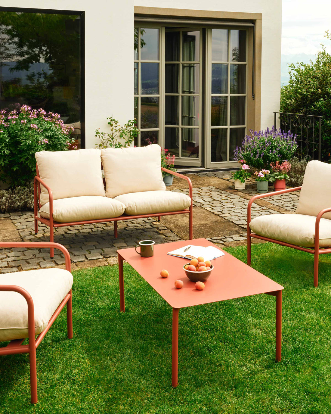 Gartenlounge mit cremefarbenen Sitzmöbeln und orangem Tisch vor einem Haus, umgeben von Blumen und Grün.