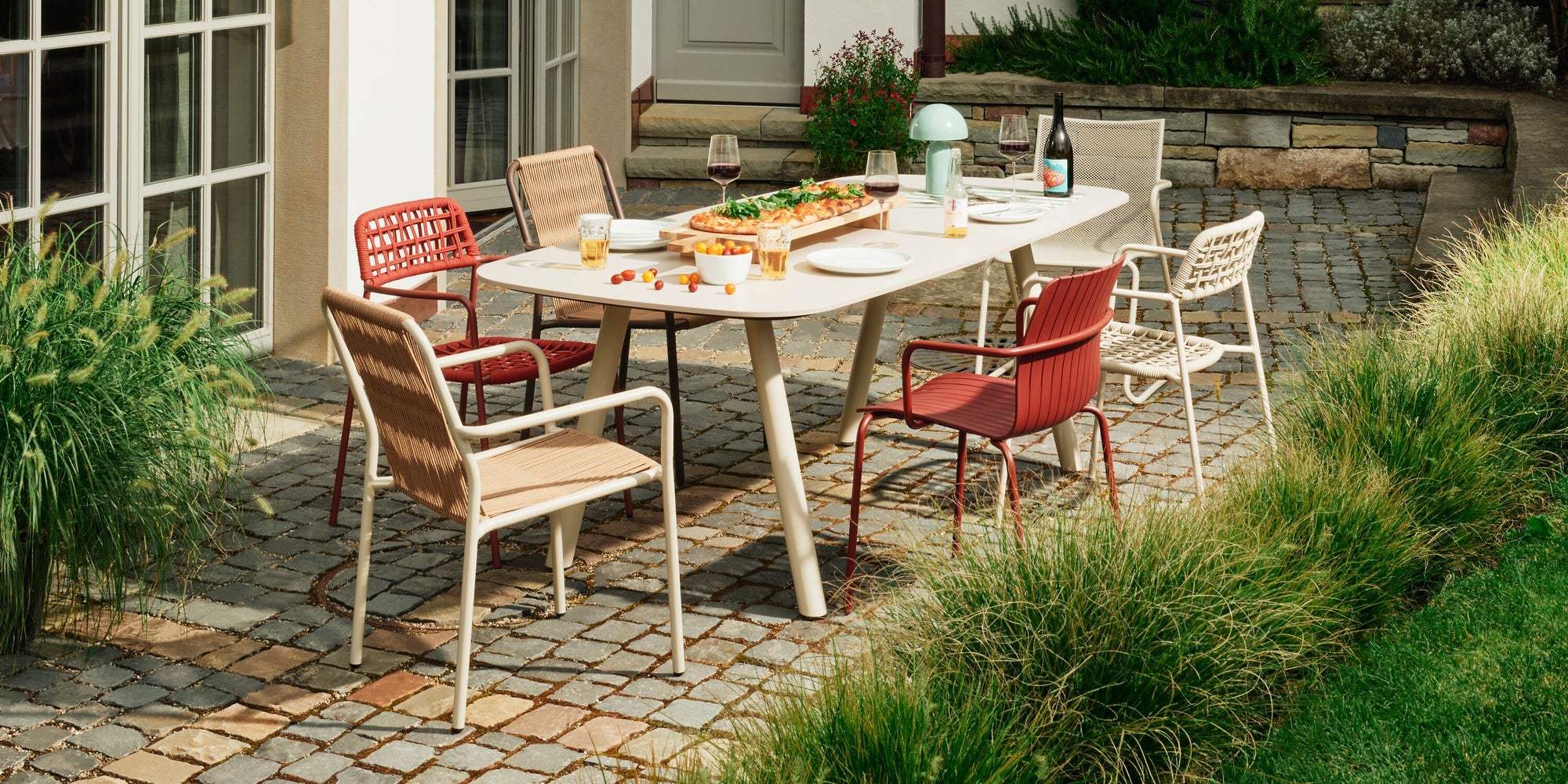 Outdoor‑Essbereich auf Steinplatte mit hellem Tisch, gemischten Gartenstühlen und sommerlich gedeckter Tafel vor einem Haus mit Terrasse.