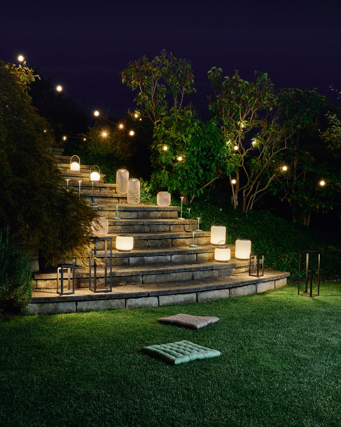 Beleuchtete Laternen und Lichterketten erhellen am Abend eine breite Steintreppe im Garten, umgeben von Bäumen und Rasen.