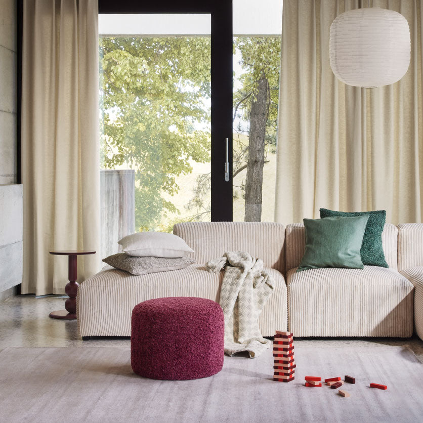 Modernes Wohnzimmer mit beigem Sofa, gruener Kissen, rotem Hocker und grossen Vorhaengen vor Fenster.