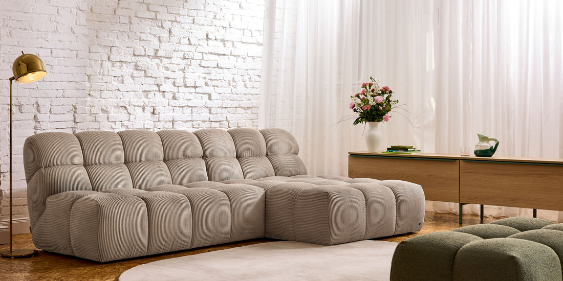 Modernes Wohnzimmer mit gepolstertem Cord-Ecksofa in Beige, Sideboard aus Holz und heller Backsteinwand.