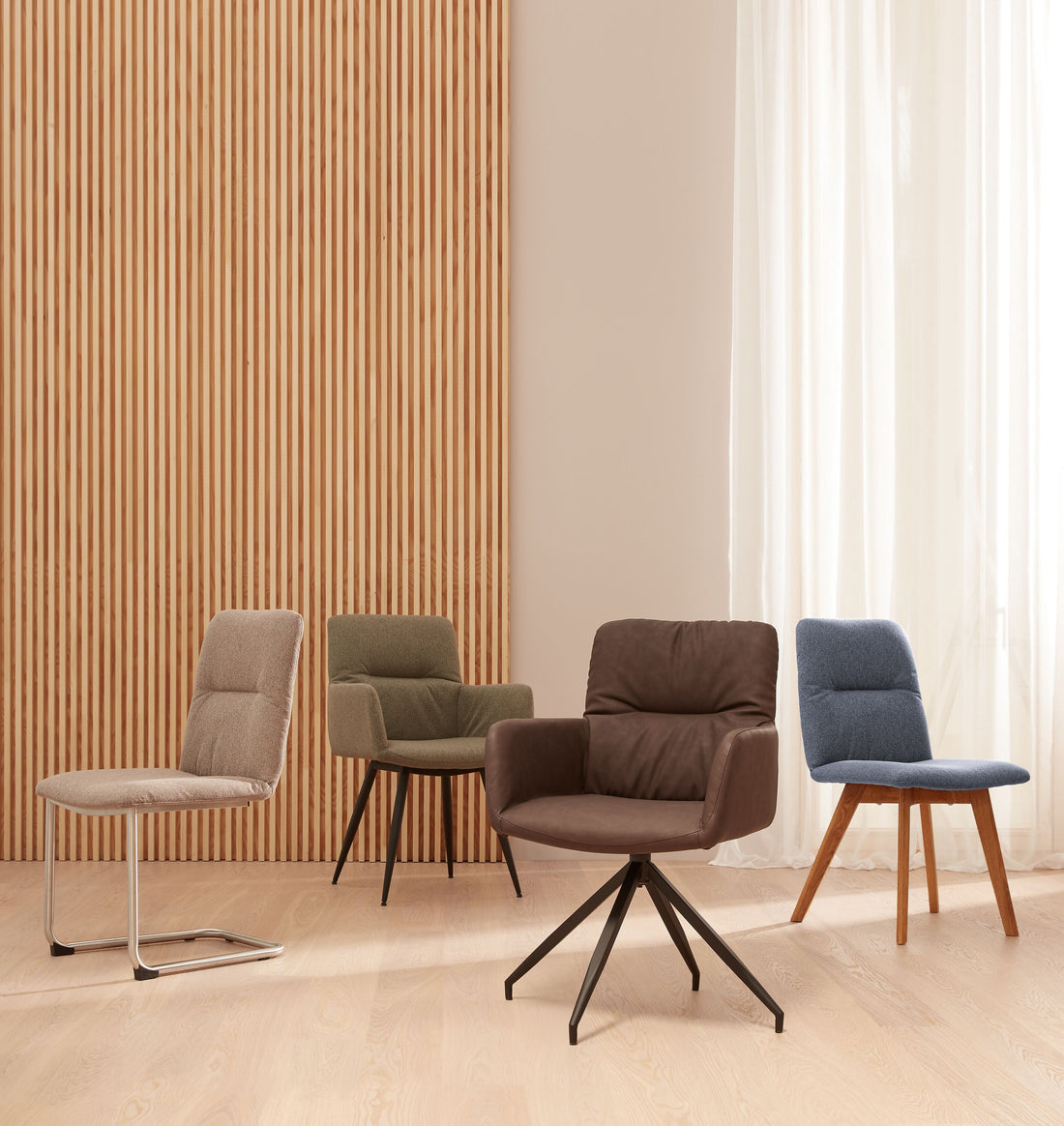 Vier moderne Polsterstühle in unterschiedlichen Farben und Gestellen stehen in einem hellen Raum mit Holzlamellenwand und großen Vorhängen.