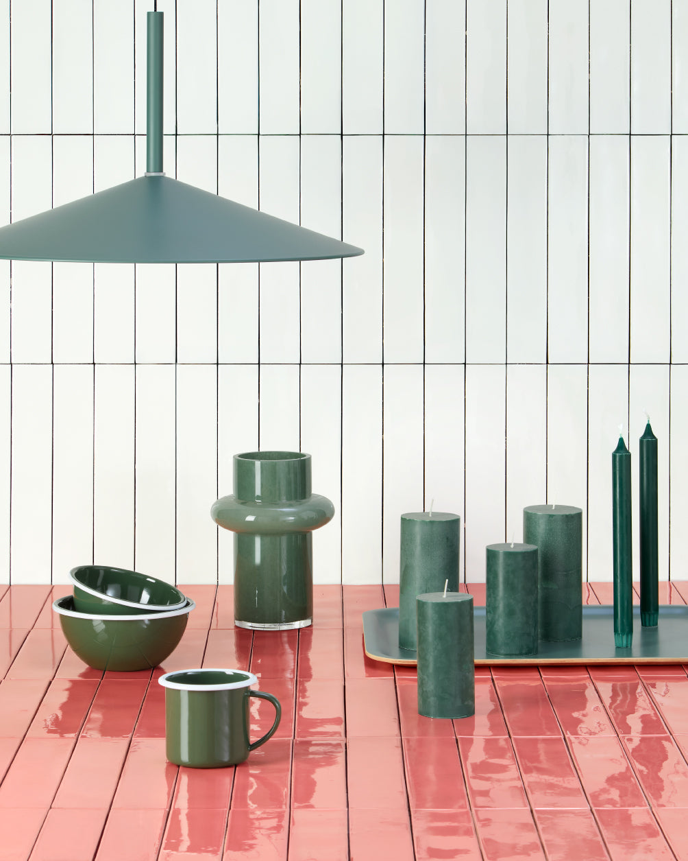 Stillleben mit grünen Kerzen, Geschirr und Vase auf rosafarbenen Kacheln unter einer modernen Hängelampe.