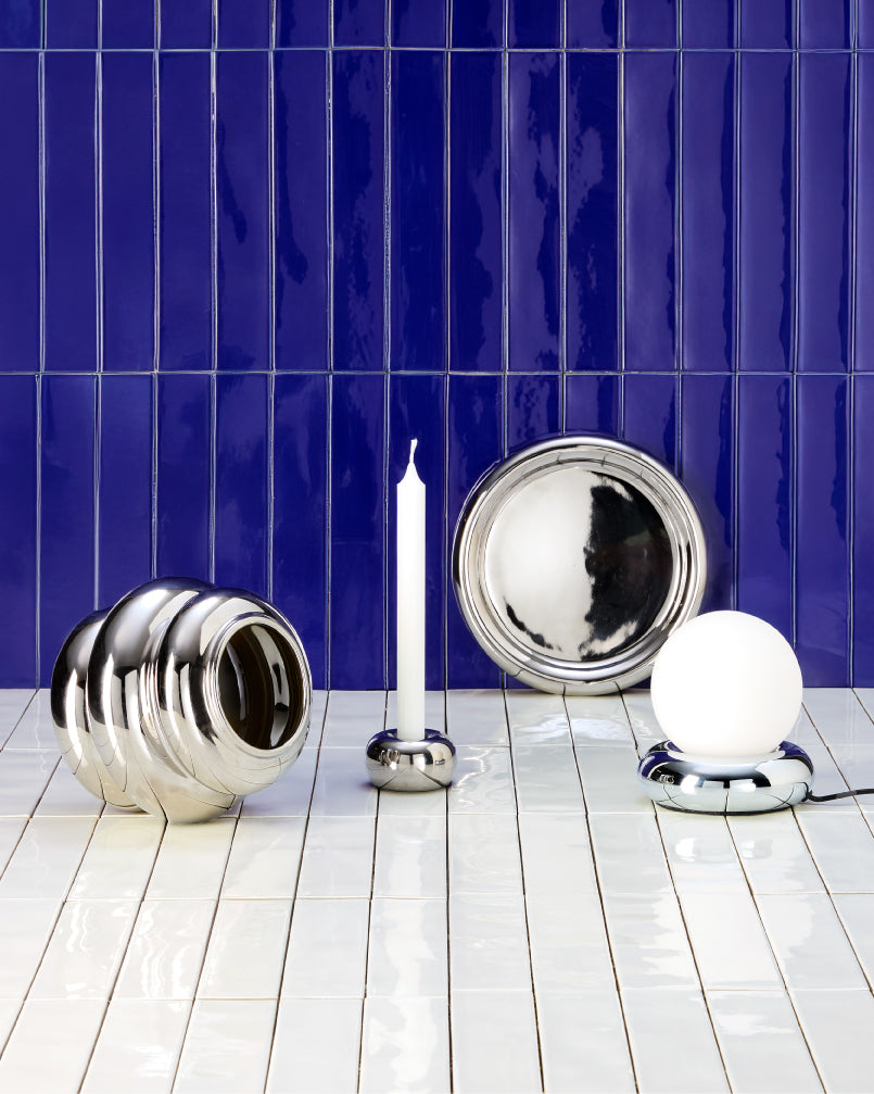 Dekoration aus silbernen Objekten, Kerzenständer und Leuchte vor dunkelblauer Kachelwand.