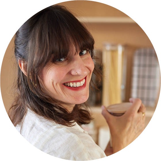 Debora mit langen Haaren hält eine Tasse und blickt freundlich in die Kamera. Im Hintergrund sind Küchenutensilien zu sehen.