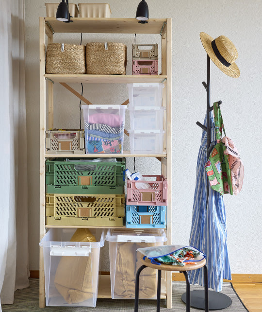 Holzregal mit bunten Aufbewahrungsboxen, daneben ein Kleiderständer mit Strohhut und gestreifter Kleidung, im Vordergrund ein Hocker mit Stoffen.
