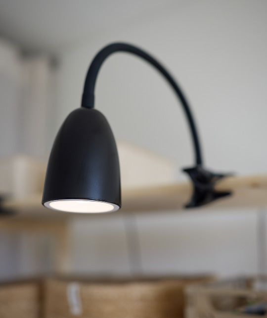 Schwarze Schreibtischlampe mit flexiblem Hals und Klemme, befestigt an einem Regal und leuchtend nach unten.