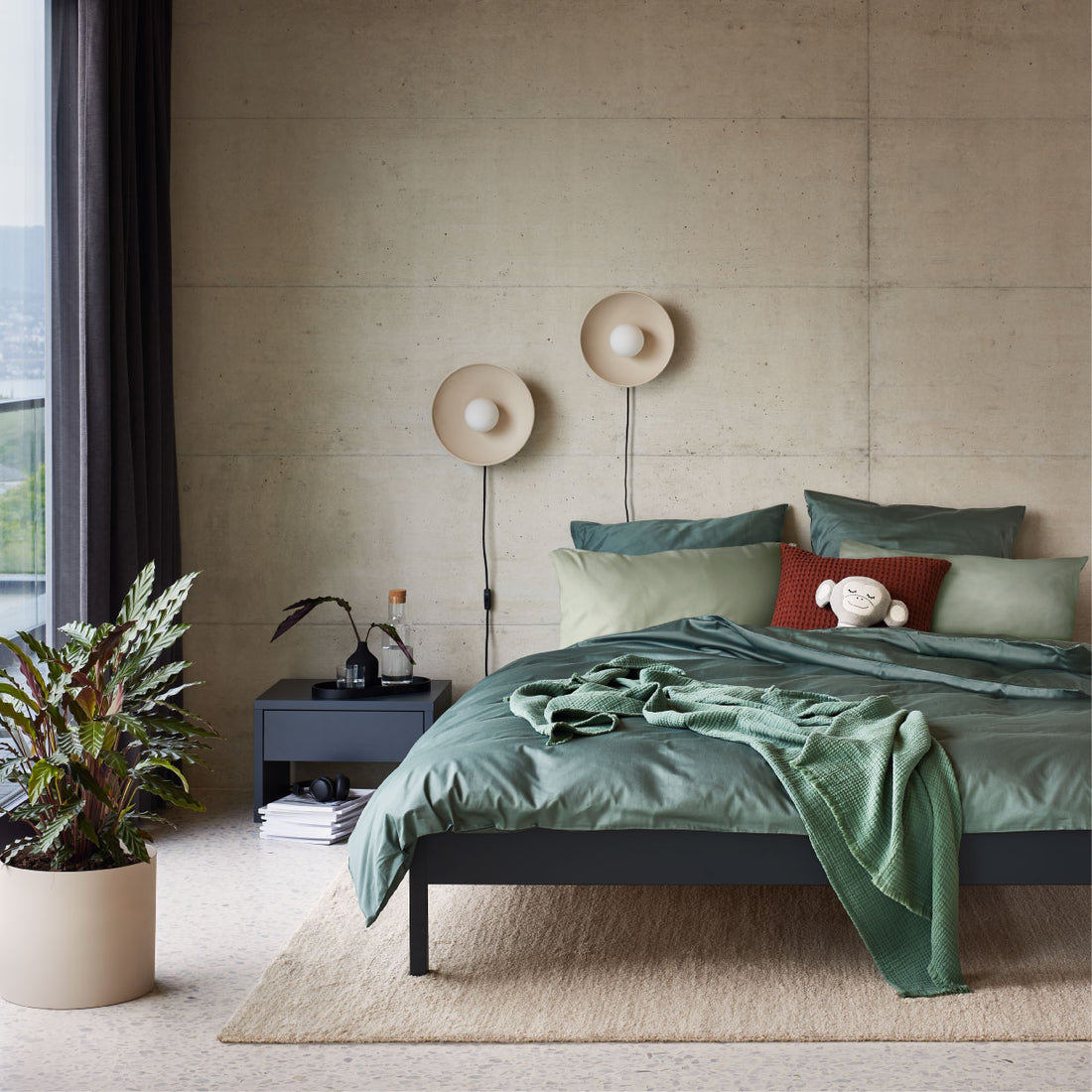 Modernes Schlafzimmer mit gruenem Bettwaesche-Set, zwei runden Wandlampen und grossem Pflanzentopf.