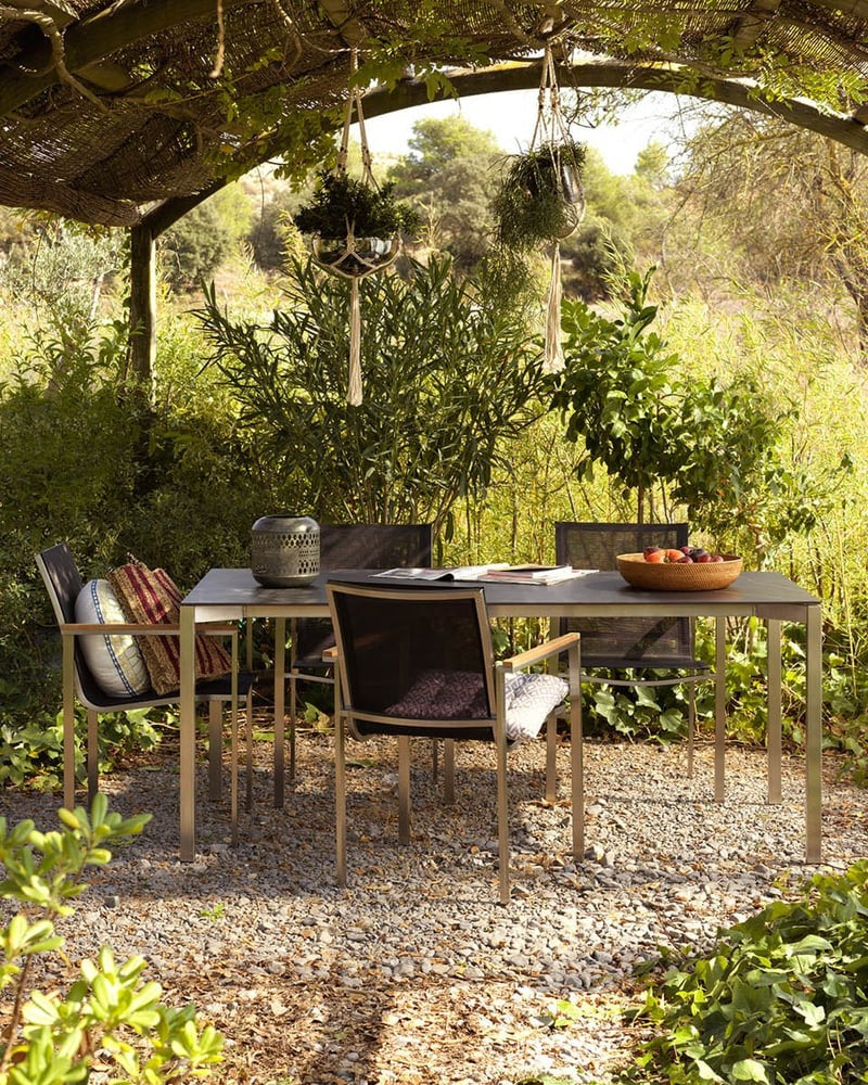 Gartenbereich mit Tisch und Stühlen unter einer Pergola, umgeben von Pflanzen.