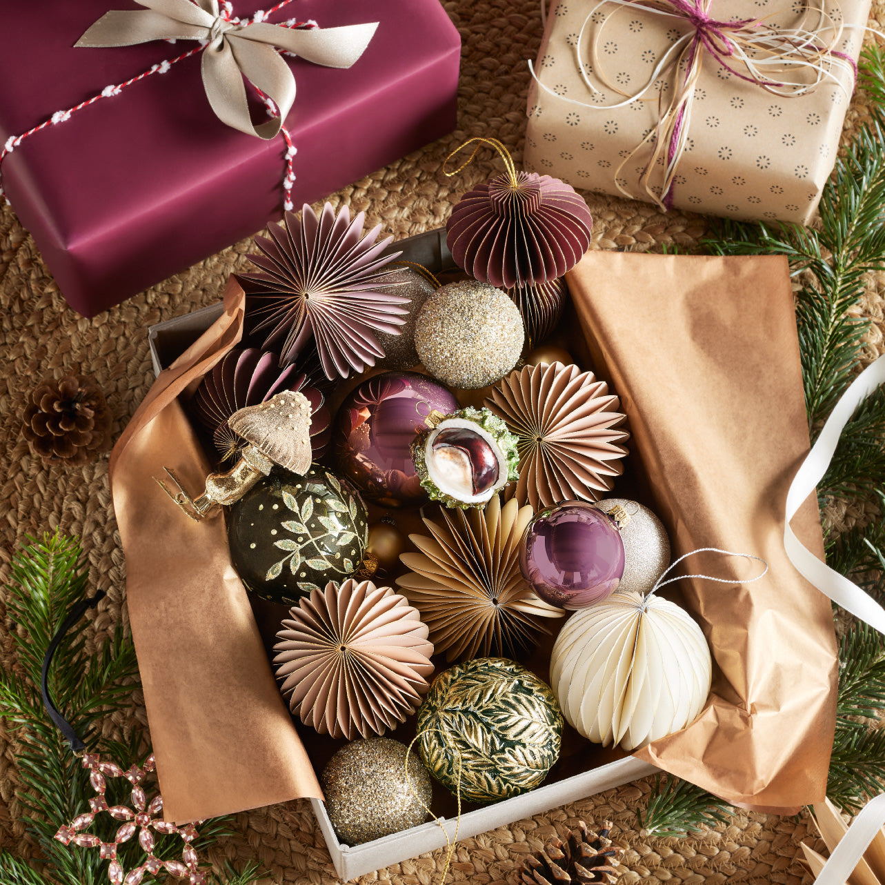 Offene Geschenkbox mit Weihnachtskugeln und Papierornamenten in Gold-, Rosa- und Violetttönen, umgeben von verpackten Geschenken und Tannenzweigen.