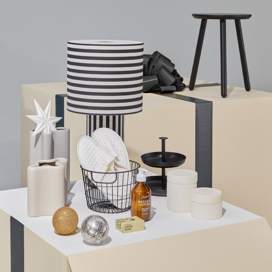 Geschenkideen für Entspannung: gestreifte Tischlampe, Hausschuhe im Drahtkorb, Lotion, Seife, Kerzen und Deko auf hellen Boxen.