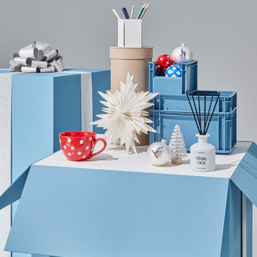 Geschenkideen für Ordnungsliebende: rote Tasse, Papierstern, Aufbewahrungsboxen, Christbaumkugeln und Duftstäbchen auf blauen Geschenkboxen.