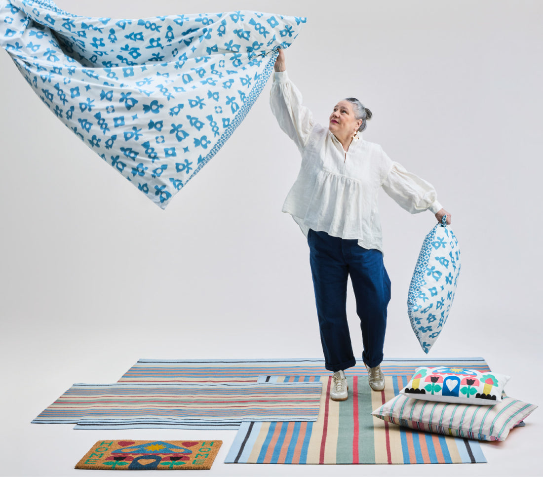Person wirft ein gemusterte Bettwäsche in die Luft, umgeben von farbigen Textilien und Teppichen im Studio‑Setting.