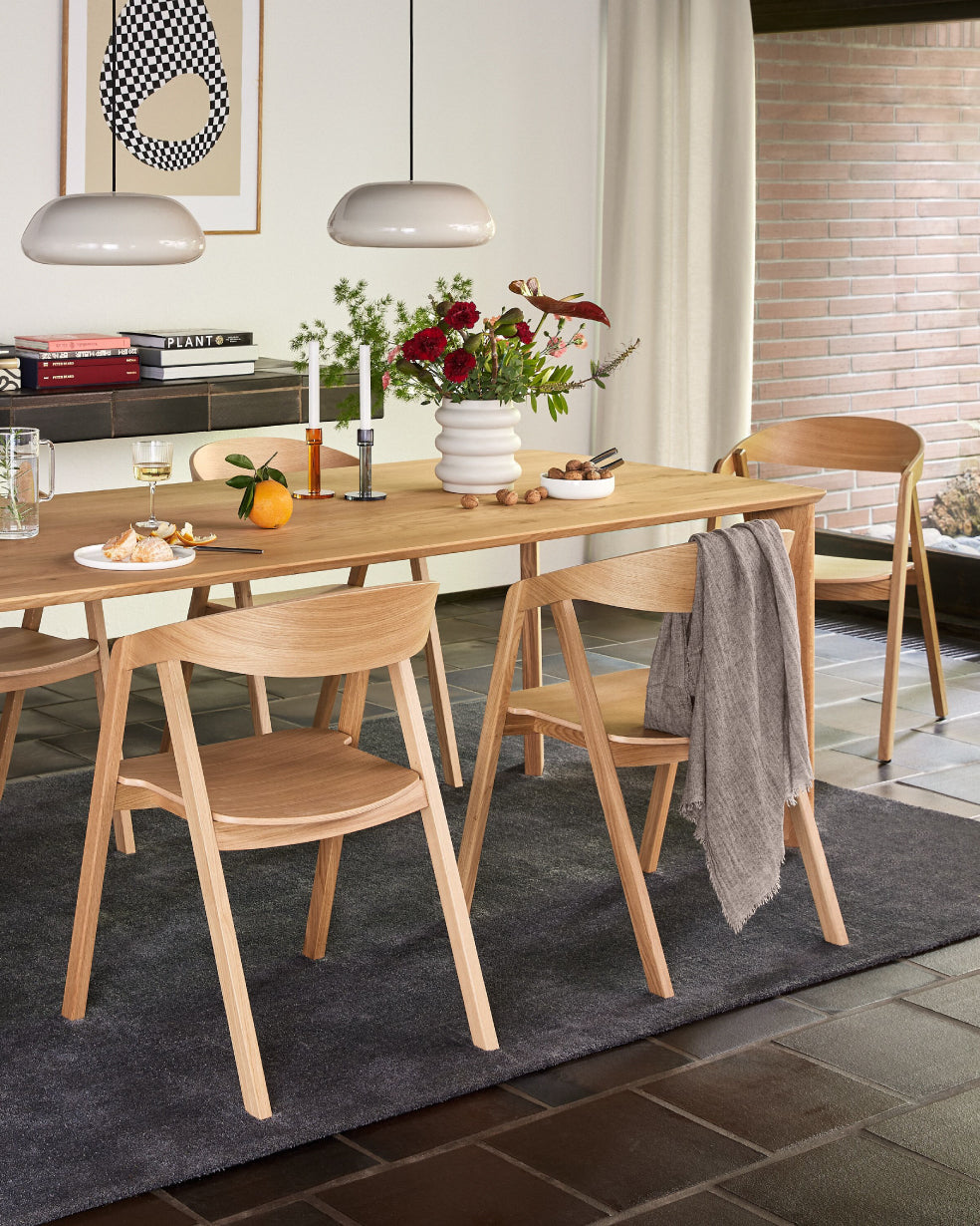 Modernes Esszimmer mit rechteckigem Holztisch, vier passenden Holzstühlen, dekoriert mit Blumen, Kerzen und Obst, unter zwei schlichten Pendelleuchten vor heller Wand mit Grafikbild.