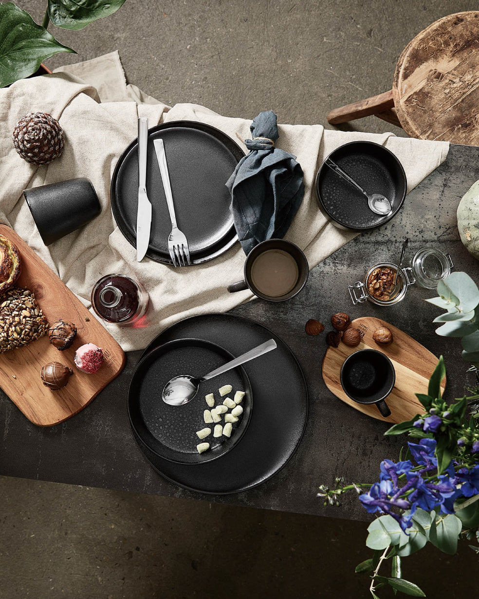 Rustikal gedeckter Tisch mit schwarzem Geschirr, Besteck, Tassen und Servietten auf einem Leinentuch, dazu Holzbretter mit Gebäck, Nüssen und einer Flasche, umgeben von dekorativen Blumen.