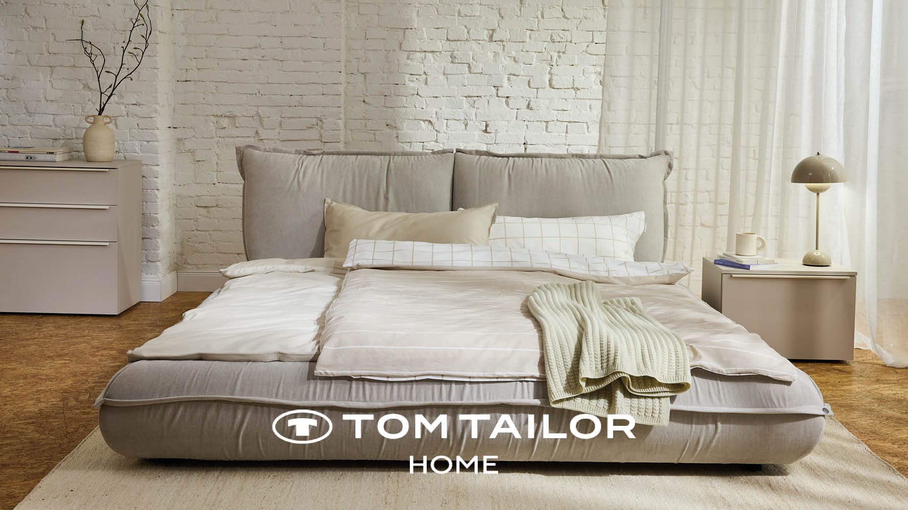 Gepolstertes Bett der TOM TAILOR HOME Kollektion in hellem Beige vor einer weissen Backsteinwand.